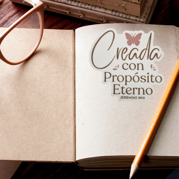 Open book with a decorative sticker Creada con propósito eterno Jeremías 29:11 on a wooden surface with glasses and a pencil.